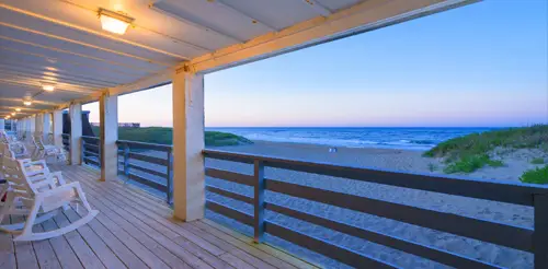 A porch with a rocking chair looking out past the dune grass to the ocean at dusk