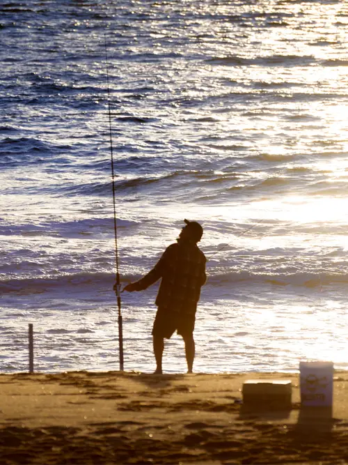 A surf fisherman silhouetted against the ocean, casting a line at dusk